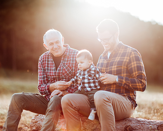 Grandpa, son and infant grandson sitting on a log outside.